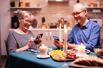 Retired man browsing on smartphone in kitchen and having dinner with wife. Sitting at the table in the kitchen, browsing, searching, using phone, internet, celebrating their anniversary in the dining