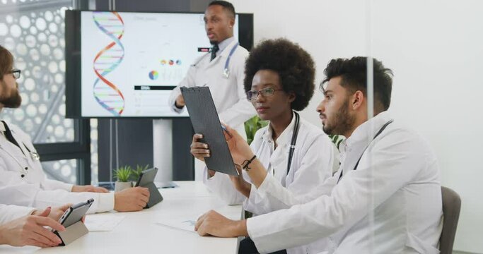 Medical concept where two multiracial confident male and female doctors discussing results of patient's examination during joint conference on dna theme with another colleagues,front view