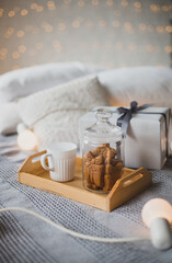 Cup with hot tea and Christmas cookies on wooden tray standing on bed in decorated room with festive lights on background.