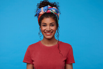 Portrait of young pretty brunette curly woman with festive makeup looking cheerfully at camera with charming smile, standing against blue background