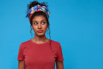 Studio photo of young lovely brown-eyed dark haired woman with orange lips looking wonderingly aside and keeping hands down while posing over blue background