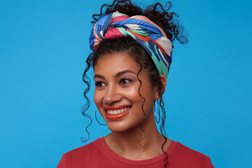 Pleased young lovely dark haired curly female with gathered hair looking happily aside with charming smile, posing over blue background with hands down