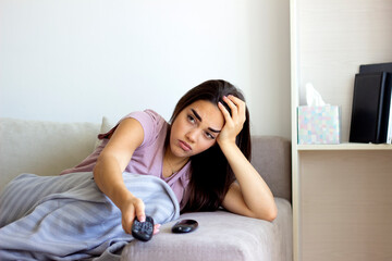 Fototapeta na wymiar Photo of a young woman sitting on a sofa, feeling depressed and changing channels on a TV remote. Young brunette woman relaxing at home, curled up in a blanket, holding the remote, watching television