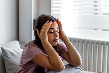Young woman suffering from headache at home. Unhappy girl suffering from headache sitting on couch in the living room. Portrait of stressed young woman with migraine. Health, stress and people concept