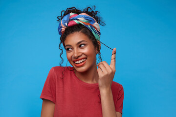 Glad young attractive dark haired female with multi-colored headband smiling happily and twisting her curly hair on finger, isolated over blue background