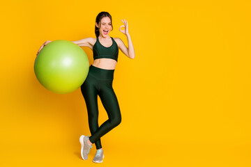 Full length body size view of nice cheerful girl holding in hands fitness ball showing ok-sign winking isolated over bright yellow color background.