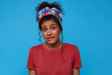 Portrait of confused young pretty curly female with gathered hair biting worringly underlip while looking perplexedly at camera, standing over blue background