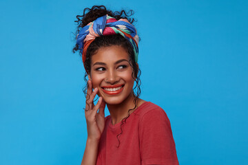 Happy young attractive brown haired curly woman with festive makeup touching her face with raised hand and smiling happily, isolated over blue background