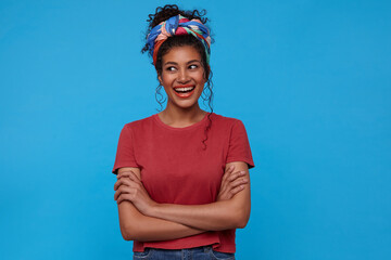 Glad young attractive dark haired curly female with festive makeup keeping hands folded while looking cheerfully aside and smiling widely, isolated over blue background