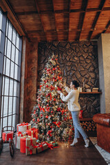 girl in a white sweater decorates a Christmas tree at home by the window - 391482748