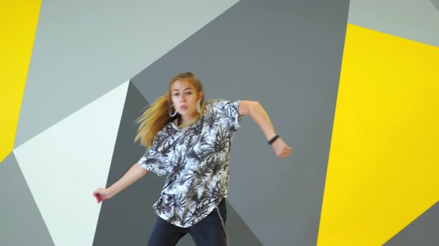 Cheerful young woman dancing hip hop in the studio in front of the wall with geometric pattern. Medium long shot