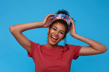 Portrait of young lovely dark haired curly female with festive makeup laughing happily and raising emotionally hands to her head, isolated over blue background