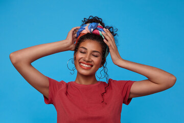 Pleasant looking young pretty brunette curly female smiling cheerfully with closed eyes and raising hands to her head, standing against blue background