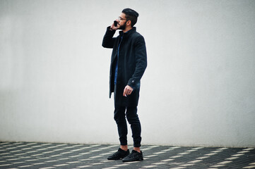 Middle eastern entrepreneur wear black coat and blue shirt, eyeglasses standing against white wall, smoking cigarette and speak by phone.