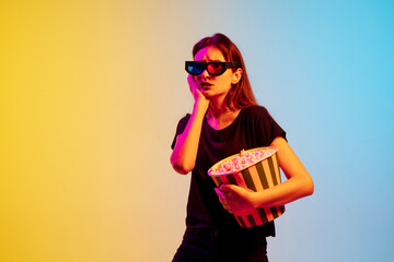 Obraz premium Sad. Young caucasian woman with bunch of popcorn on gradient blue-yellow studio background in neon. Concept of movie, human emotions, facial expression, sales, ad. Watching cinema in eyewear.
