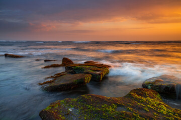 Beautiful seascape. Beach with stones covered by seaweeds. Low tide. Composition of nature. Motion water. Cloudy sky with sunlight. Slow shutter speed. Mengening beach, Bali, Indonesia