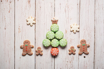 Gingerbread family standing by a Christmas tree made of macarons with a gingerbread star topper and snowflake cookies falling on a weather white table background