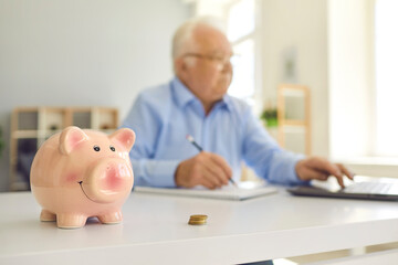 Piggy bank standing on desk with blurred retired man using laptop and doing accounting in background - obrazy, fototapety, plakaty