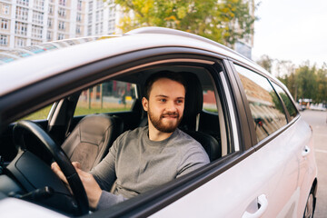 Happy bearded young man sitting in car and using cell phone, side view, looking away. Handsome businessman using mobile phone in auto. M