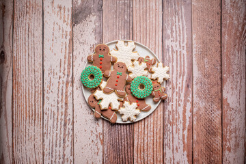Christmas cookies and gingerbread man cookies on a full white plate.  Icing and decorations on the gingerbread men, wreaths and snowflakes on a weathered wooden table background with copy space.