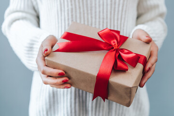 Close up cropped shot of woman hands with red polished nails holding gift box with red satin ribbon bow