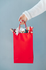 Cropped image of female hand with red polished nails holding plain red shopping bag full of christmas gift boxes