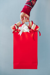 Cropped image of female hand with red polished nails holding plain red shopping bag full of christmas gift boxes