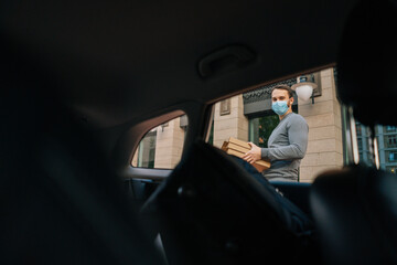 Shooting from interior car of delivery man wearing medical protective mask standing at city street and holding carton boxes with hot pizza. Concept of safe food delivery service during quarantine.