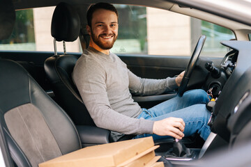 Cheerful delivery man wearing casual clothes driving car delivered hot pizza to customer, looking at the camera. Concept of fast online delivery around the city.