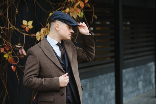 A man posing in the image of an English retro gangster of the 1920s dressed in Peaky blinders style near old brick wall.