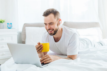 Portrait of a smiling young man in pajamas using a laptop, listening to music and drinking coffee while lying in bed in the morning chilling in the hotel at the vacation