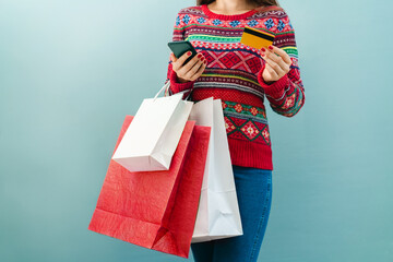 Cropped shot of female customer wearing cozy knitted red sweater with Christmas ornament and holding bunch of shopping bags, moblile phone and credit card in hands