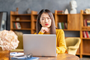 Unhappy frowning asian woman forgetting smth working on laptop at home or in cafe. Young lady in bright yellow jumper is sitting at desk typing on computer
