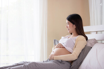 Young beautiful pregnant woman touching belly near the window in bedroom. Pregnancy, motherhood, people and expectation concept.