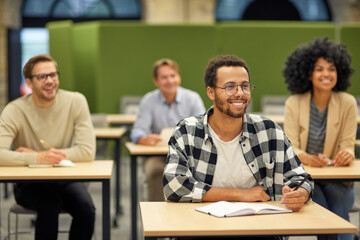 Young cheerful mixed race male office worker sitting listening to coach during seminar or training and smiling while studying with colleagues in coworking space