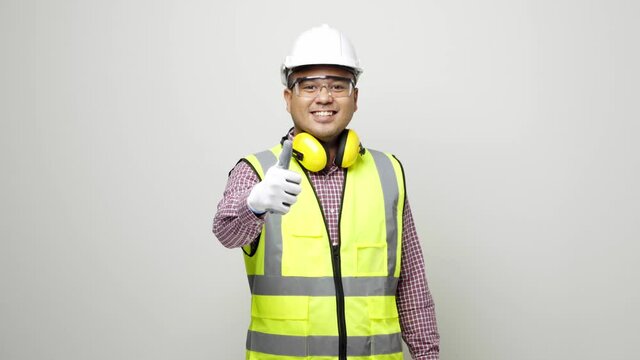 Smiling young asian civil engineer in uniform earmuff helmet safety glasses standing and showing thumbs up. 4k resolution.