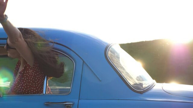 Young woman looking out window of moving vintage auto on sunny day. Happy girl in sunglasses leaning out of retro car window and enjoying trip. Travel and freedom concept. Slow motion Close up