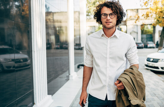 Handsome Businessman In Eyewear Smiling Broadly Posing Outdoors. Male Entrepreneur Resting In The City Street. Smart Guy In Casual Wears Spectacles With Curly Hair Walking Outside After Work