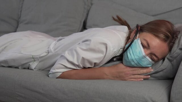 A tired and sleepy woman doctor in a medical mask falls on the couch and falls asleep. In a dream, a woman removes the mask from her face to make it easier to breathe.