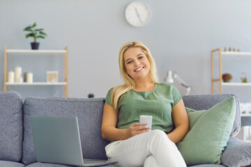 Happy woman sitting on couch with laptop, holding mobile phone in hand and looking at camera