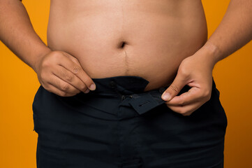 Naklejka premium Close up of overweight man trying to wear too small jeans isolated on yellow background.