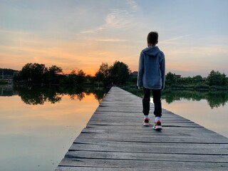 A boy walks along a wooden pier on the lake. A child is standing on the fishing platform. The guy walks across the bridge on the river.