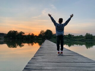 Boy with raised hands on the lake. A child is standing on a wooden pier. A young man on the shore of a reservoir.