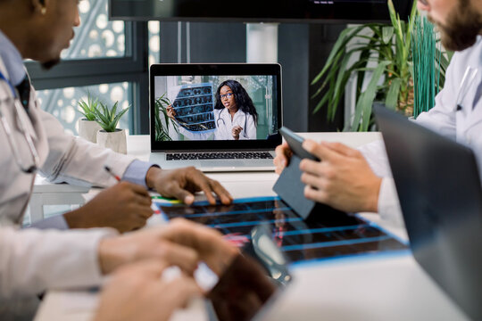 Cropped image of multiracial doctors using laptop for video conference with African female colleague, discussing CT scan results and making notes on tablets. Focus on African woman on laptop screen