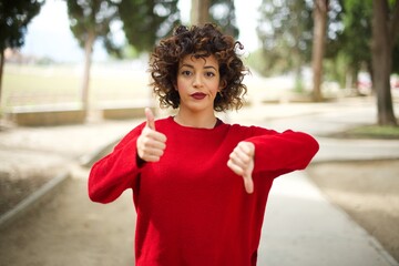 Young arab woman wearing casual red sweater in the street showing thumbs up and thumbs down, difficult choose concept - obrazy, fototapety, plakaty