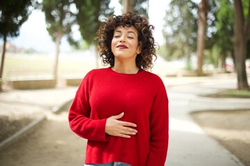 Young arab woman wearing casual red sweater in the street touches tummy, smiles gently, eating and...