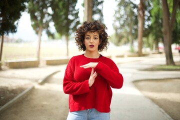 Young arab woman wearing casual red sweater in the street being upset showing a timeout gesture, needs stop, asks time for rest after hard work, demonstrates break hand sign