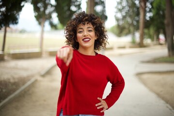 Young arab woman wearing casual red sweater in the street, pointing at camera with a satisfied, confident, friendly smile, choosing you