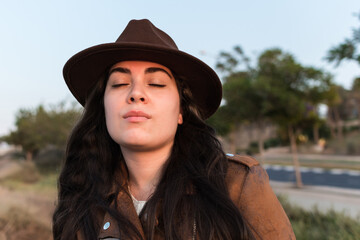 Young woman in hat and brown leather jacket, with closed eyes, in natural park.