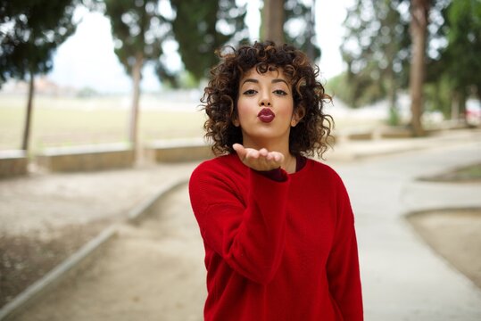Young beautiful Arab woman standing outdoors wearing red sweater looking at the camera blowing a kiss with hand on air being lovely and sexy. Love expression.
